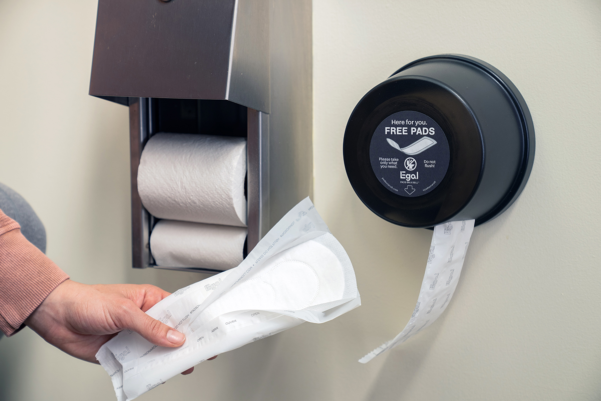 A hand taking a free pad from the Egal Pads on a Roll dispenser mounted beside the toilet paper